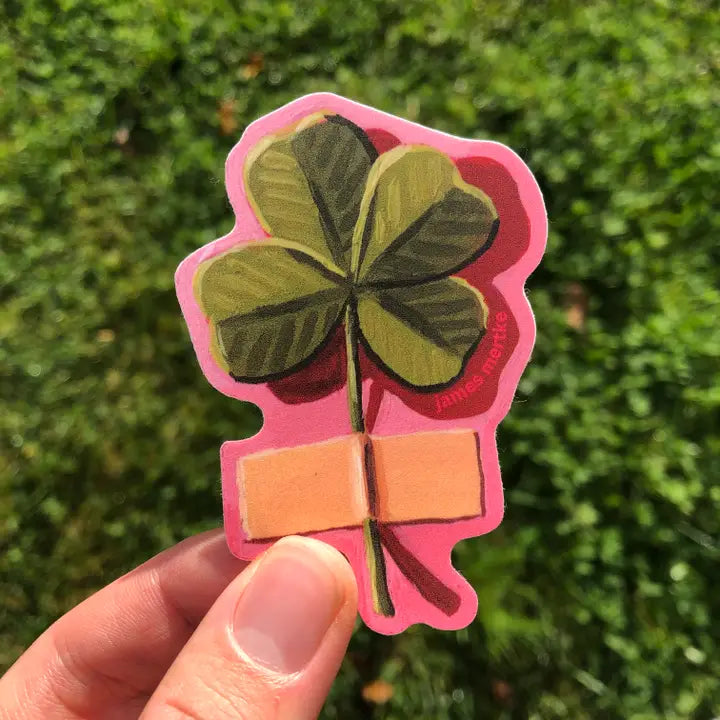 This sticker depicts a painting of a 4 leaf clover stick to the wall with a piece of tape by its stem. It has a pink outline.