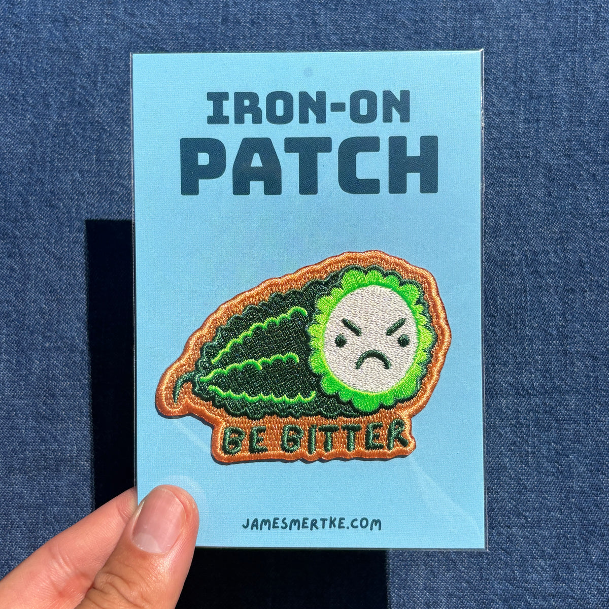An embroidered iron-on patch featuring a green bitter melon with an angry face and the text 'BE BITTER' on a light blue backing card, held by a hand against a blue denim background.