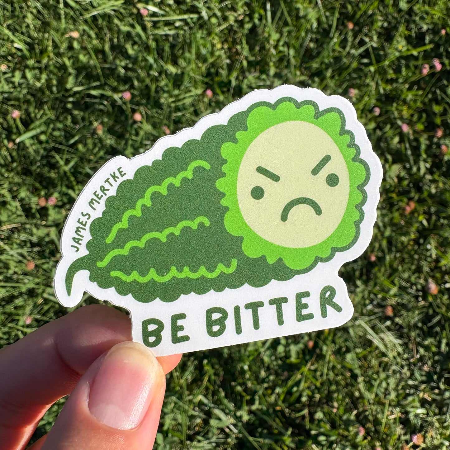 This sticker features an illustrated piece of bitter melon with a grumpy face on its cross section. The phrase "BE BITTER" is written along the bottom. It is held in a hand.