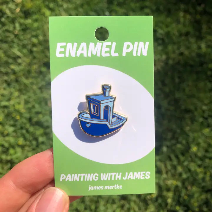 A blue boat enamel pin is attached to a green backing card. The boat is inspired by "Benchy", a common calibration print used to set up 3D printers.