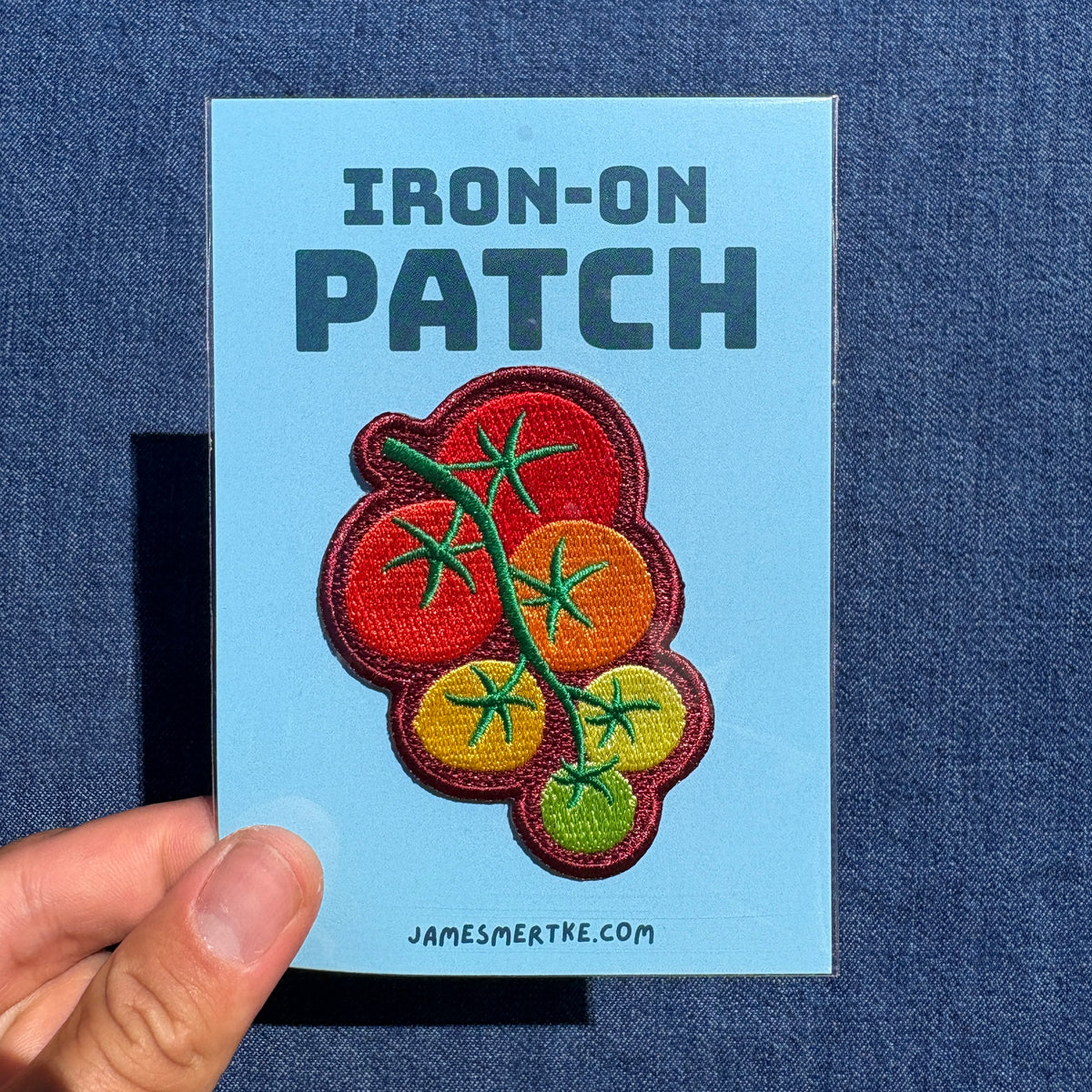 A hand holds a light blue backing card displaying an iron-on patch. The patch features embroidered cherry tomatoes on a vine in red, orange, yellow, and green.