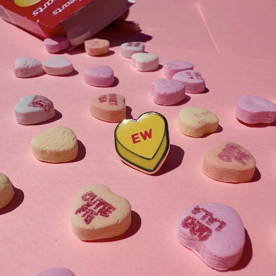 This enamel pin depicts a yellow candy conversation heart with "EW" written on the front in red text.
It is staged with candy hearts and a candy box as props.