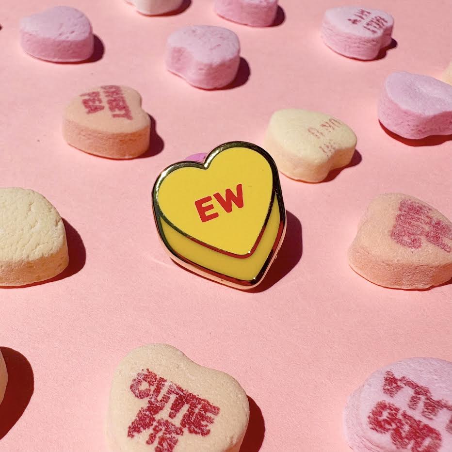 This enamel pin depicts a yellow candy conversation heart with "EW" written on the front in red text.
It is staged with candy hearts as props.