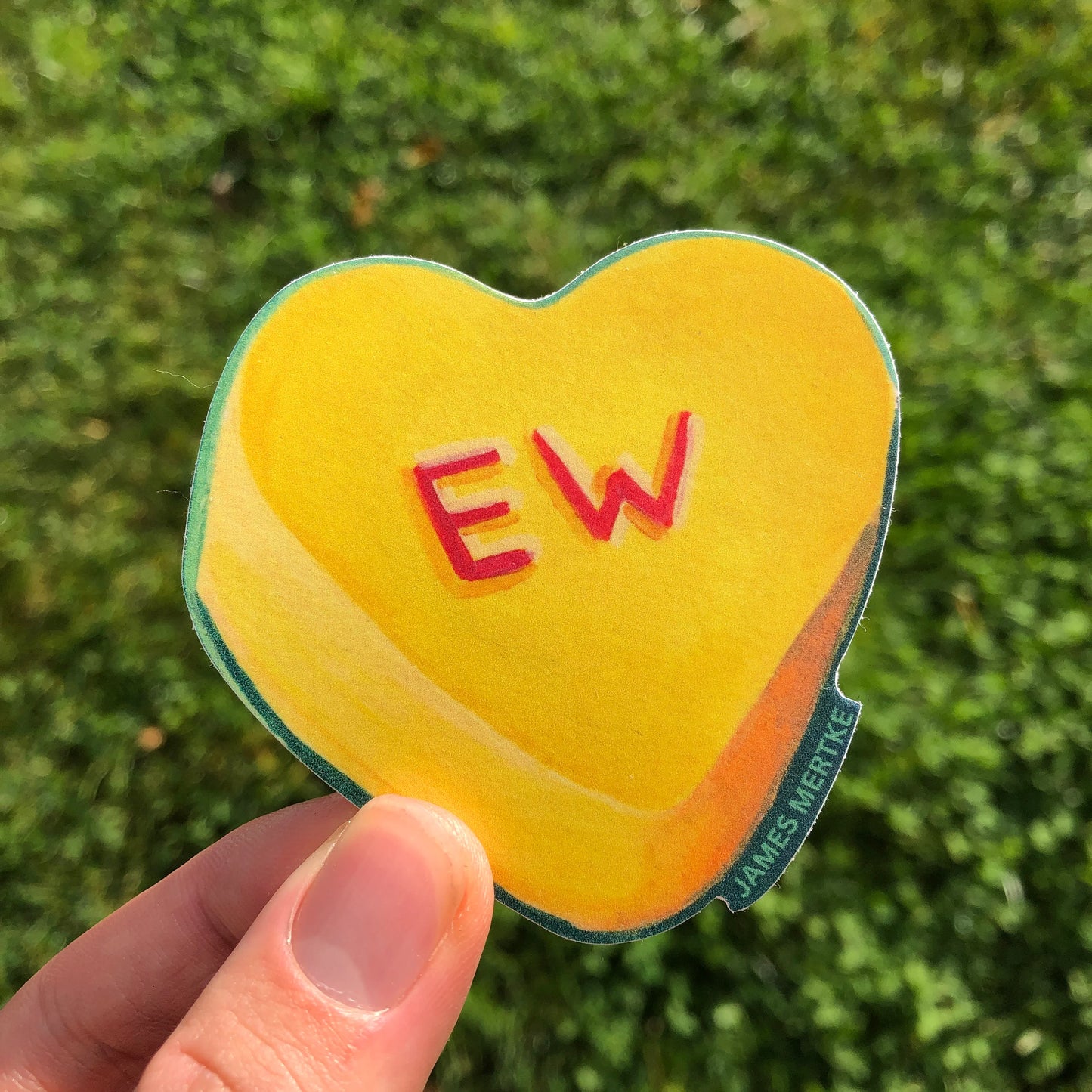 This sticker features a painting of a yellow conversation heart candy with "EW" written in red on the front.