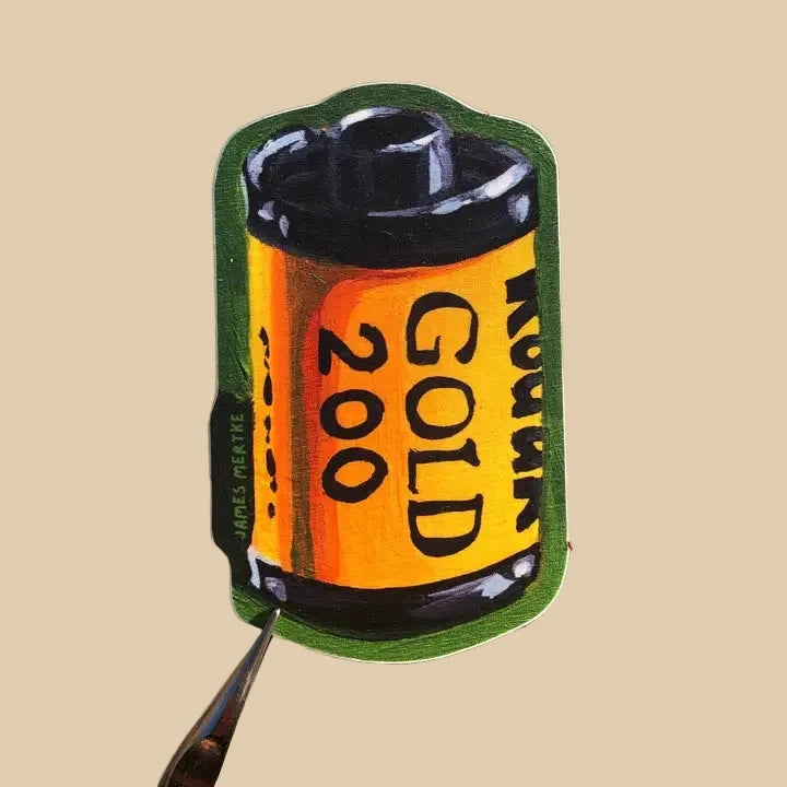This sticker depicts a painting of an analog film roll. It has a yellow base with the phrase "GOLD 400" written on its side. The background is green.