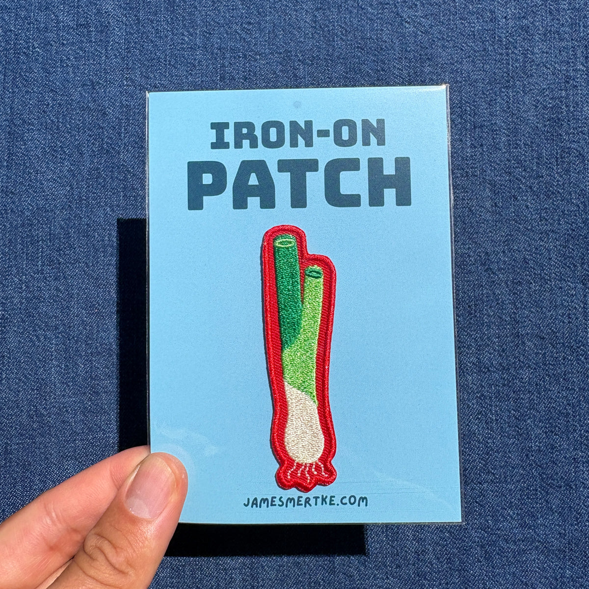 A hand holds a light blue backing card featuring an embroidered patch design of a green onion with the text 'IRON-ON PATCH' and 'JAMESMERTKE.COM'.