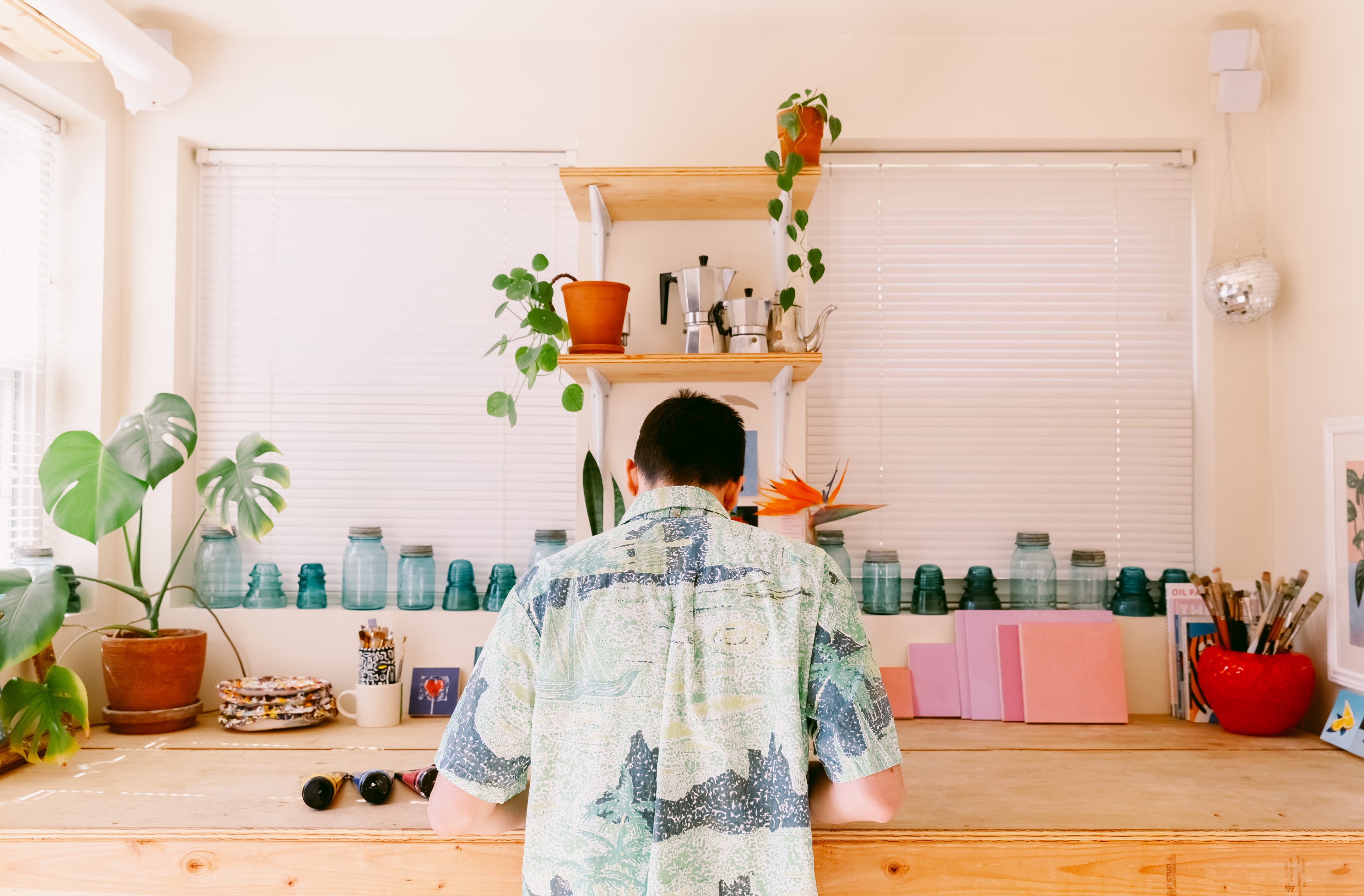James Mertke is working in his studio. He is centered in the middle of his desk and his back is facing the camera. Plants, glass jars, and art supplies cover the table.