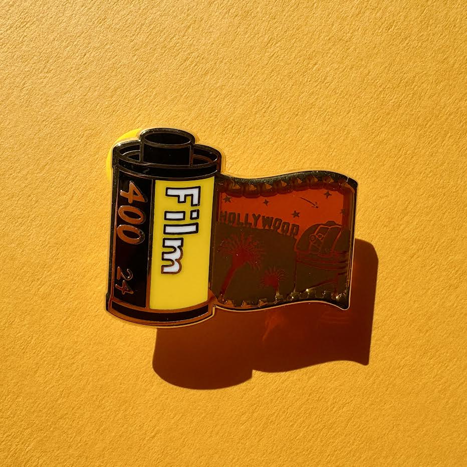 This pin features a film roll canister with "Film 400 24" written on it. It features a "stained glass" transparent panel with a screen-printed detail of Los Angeles including the Hollywood sign, Griffith Observatory, and palm trees when held up to the light.
