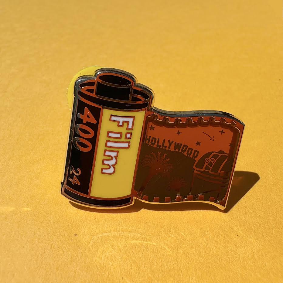 This pin features a film roll canister with "Film 400 24" written on it. It features a "stained glass" transparent panel with a screen-printed detail of Los Angeles including the Hollywood sign, Griffith Observatory, and palm trees when held up to the light.