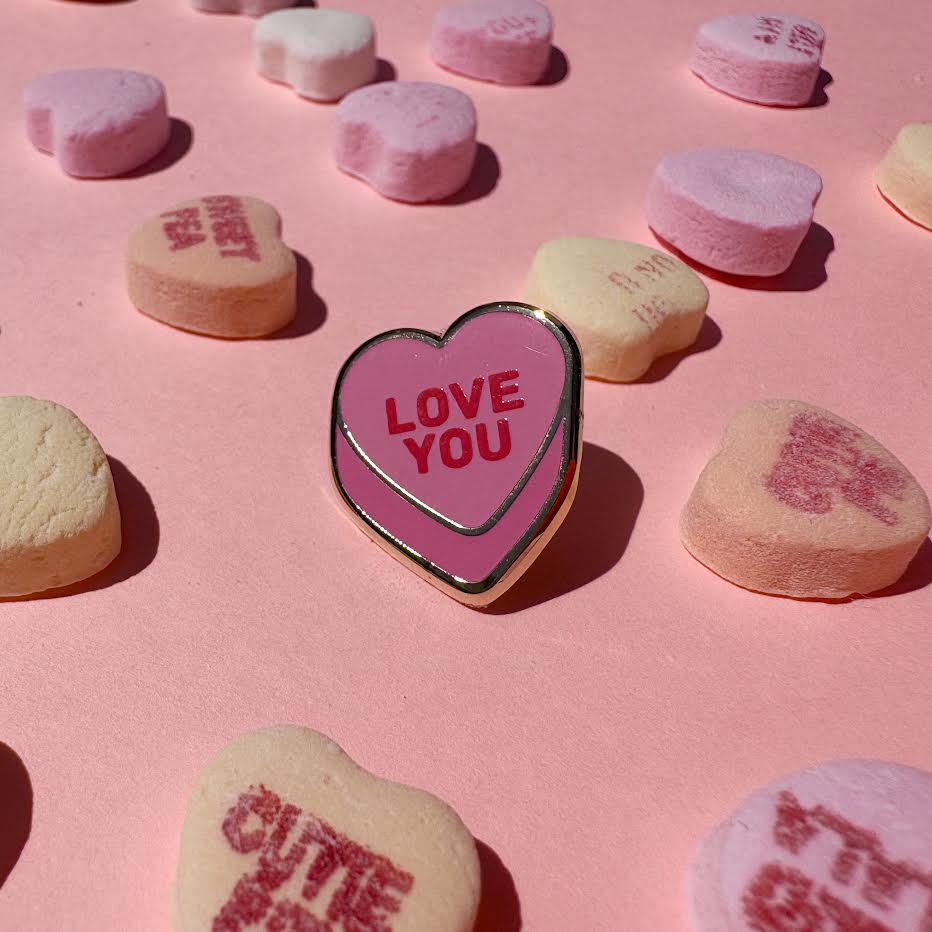 This enamel pin depicts a pink candy conversation heart with "LOVE YOU" written on the front in red text.
It is staged with candy hearts as props.