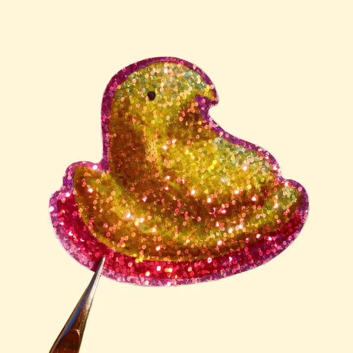 This sticker depicts a yellow marshmallow peeps treat with a pink outline. The sticker is made with a special material to give it a glittery and sparkly effect.