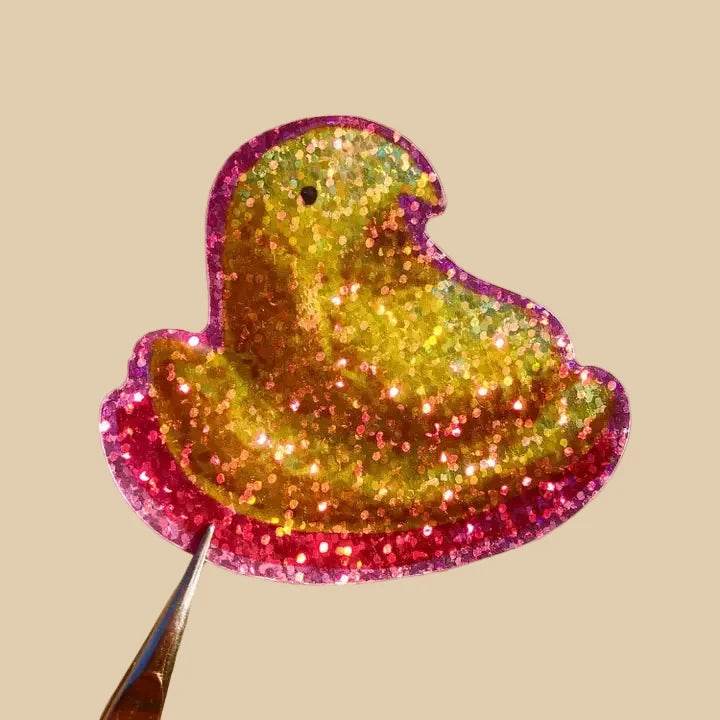 This sticker depicts a yellow marshmallow peeps treat with a pink outline. The sticker is made with a special material to give it a glittery and sparkly effect.