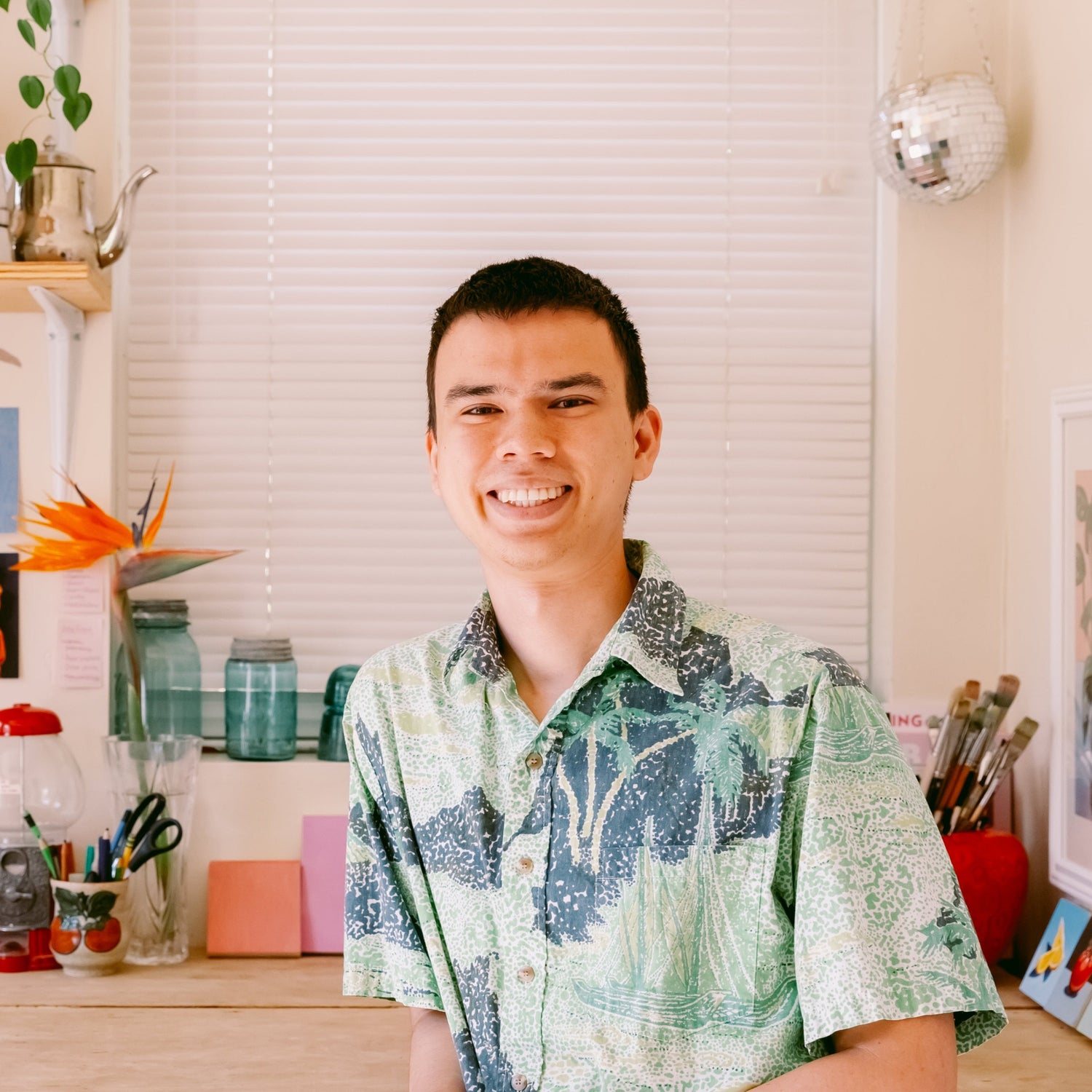A portrait of James Mertke from the waist up in front his work desk. In the background art supplies, plants, art prints, and a disco ball can be seen.