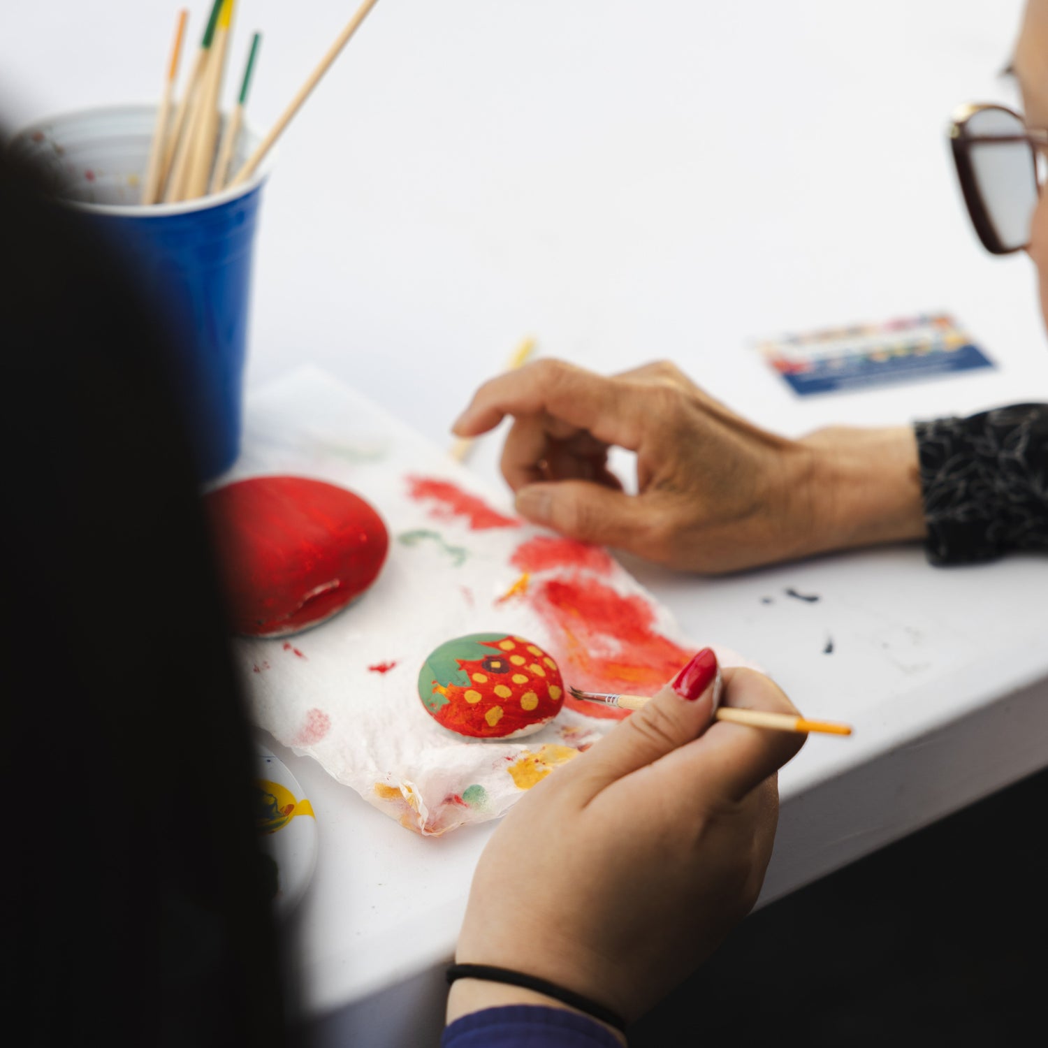An over the shoulder shot shows a woman's hand painting a round rock to look like a strawberry.