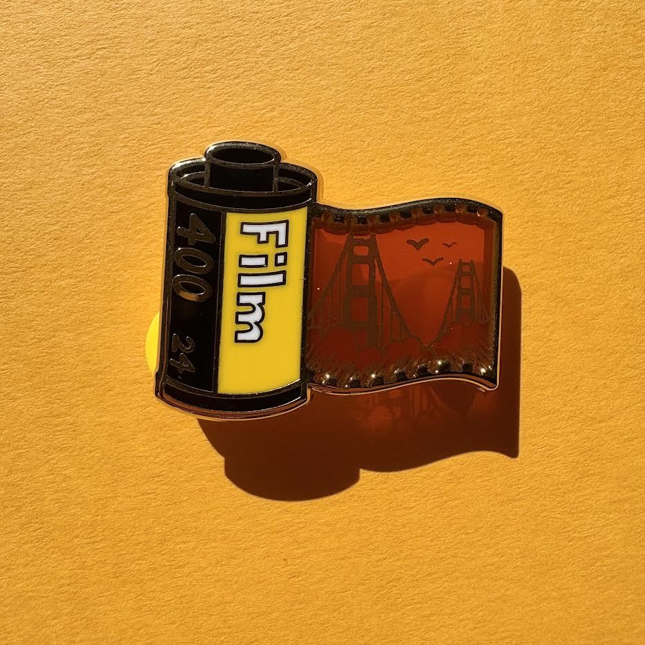 This pin features a film roll canister with "Film 400 24" written on it. It features a "stained glass" transparent panel with a screen-printed detail of San Francisco and the Golden Gate bridge when held up to the light.