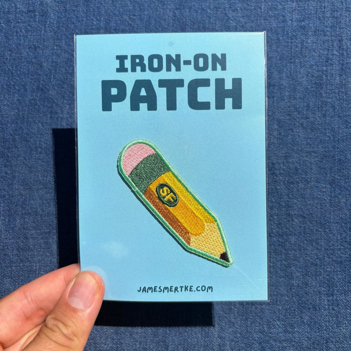 A hand holds a light blue backing card displaying an embroidered iron-on patch shaped like a yellow pencil with pink, green, and orange details. The card reads 'IRON-ON PATCH' and 'JAMESMERTKE.COM'