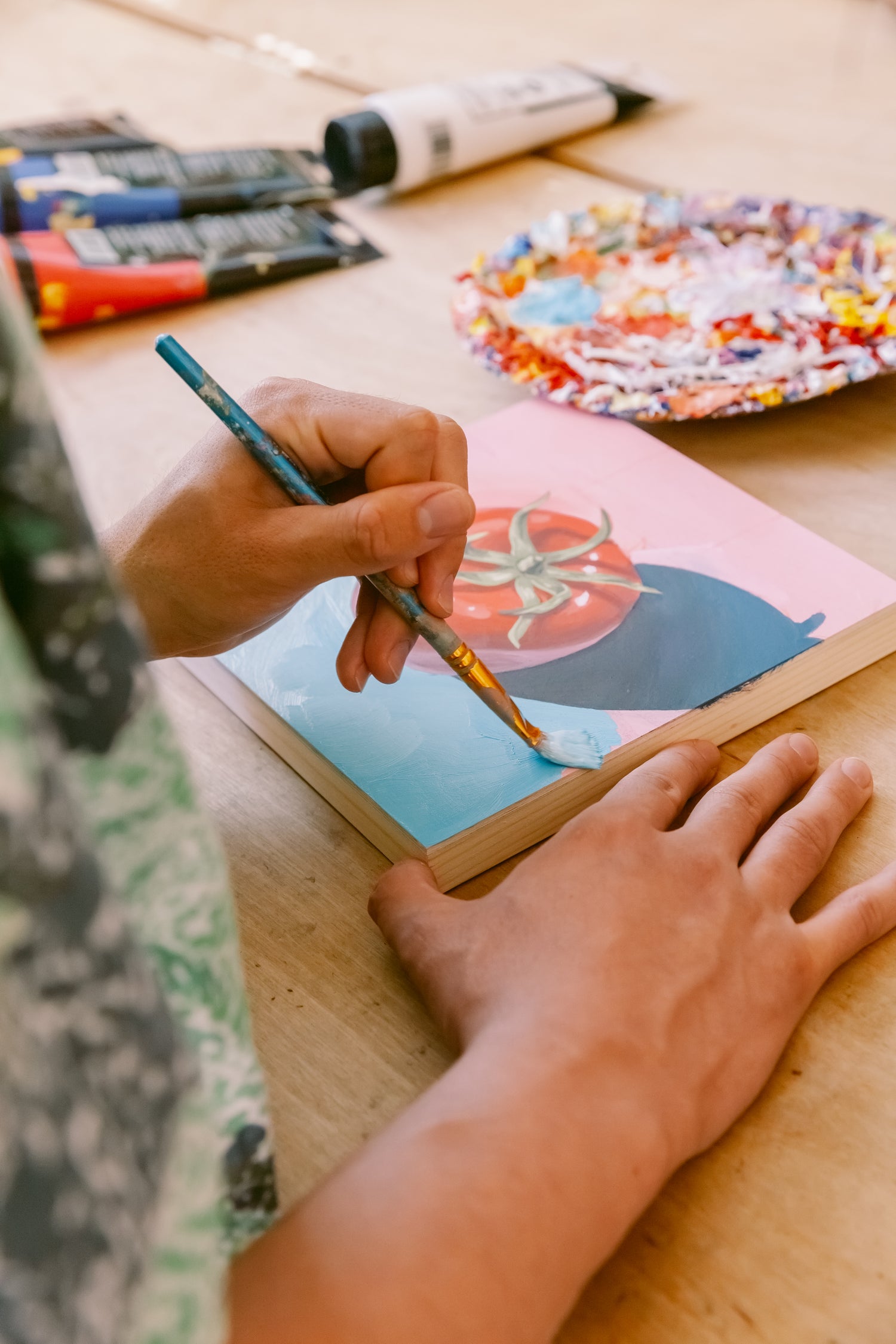 A side shot showing James painting the blue background of a tomato painting. The brush is in his left hand and his right hand stabilizes the panel in the center.
