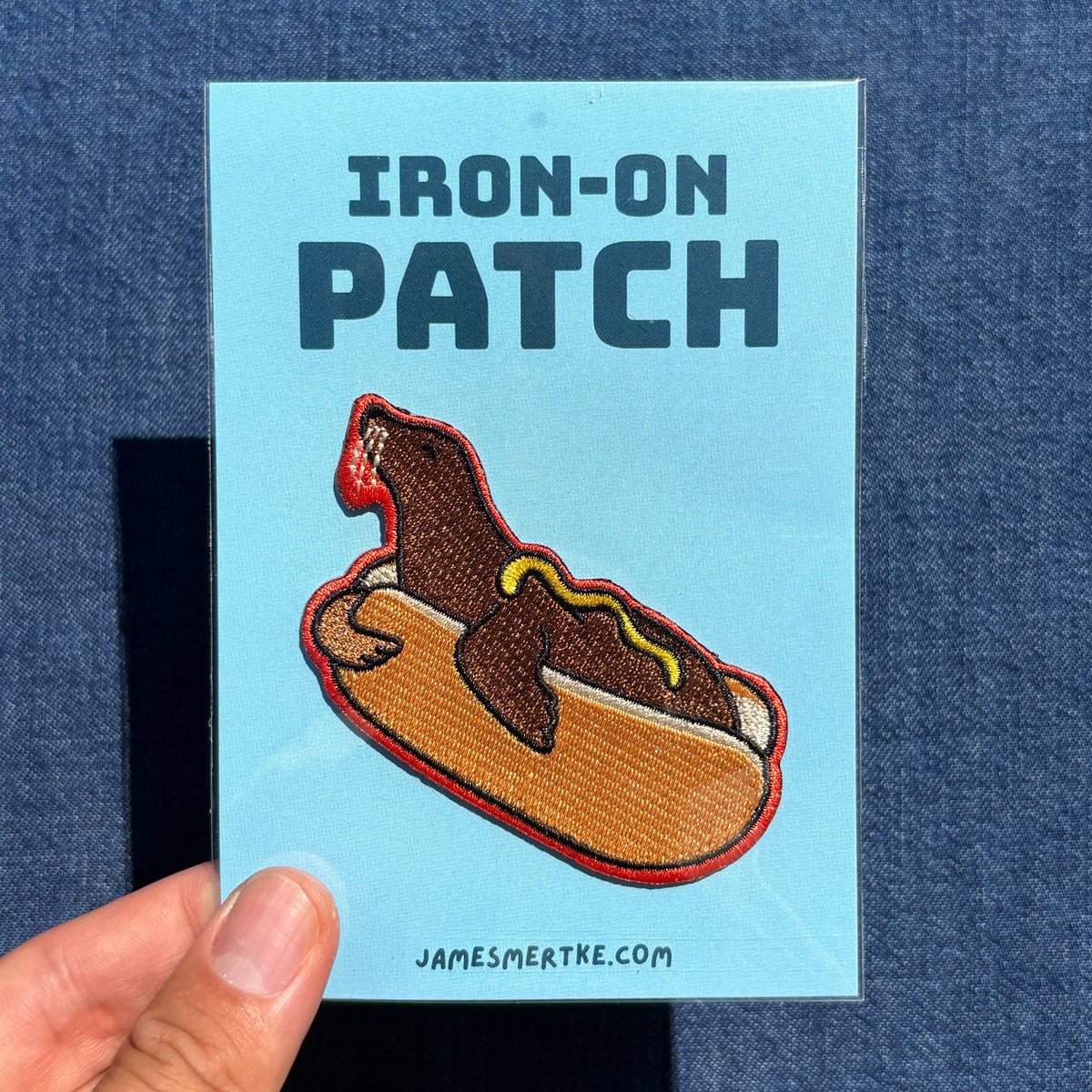 A hand holds a light blue card labeled 'IRON-ON PATCH' featuring an embroidered sea lion hot dog patch. The card also shows the website JAMESMERTKE.COM .