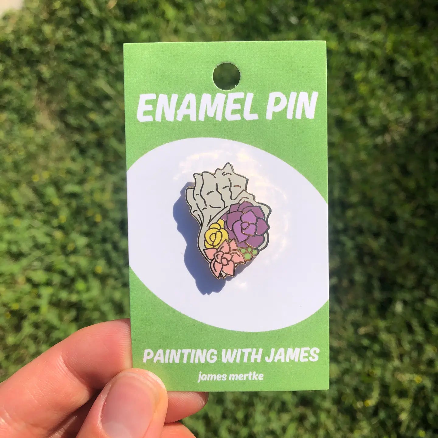 An enamel pin depicts succulents growing out of the opening of a conch shell. The succulents are purple, pink, yellow, and green in color.