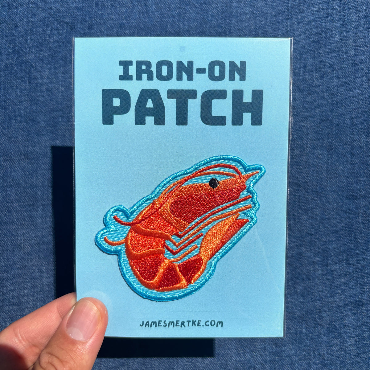 A hand holds a light blue backing card featuring an embroidered iron-on patch of a stylized orange shrimp with a blue outline. The card reads 'IRON-ON PATCH' and 'JAMESMERTKE.COM'