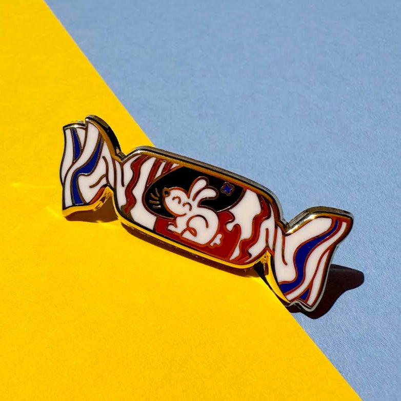 This enamel pin depicts a white rabbit candy with its red, white, and blue wrapper with a cheerful bunny. The pin is displayed on a yellow and blue background.