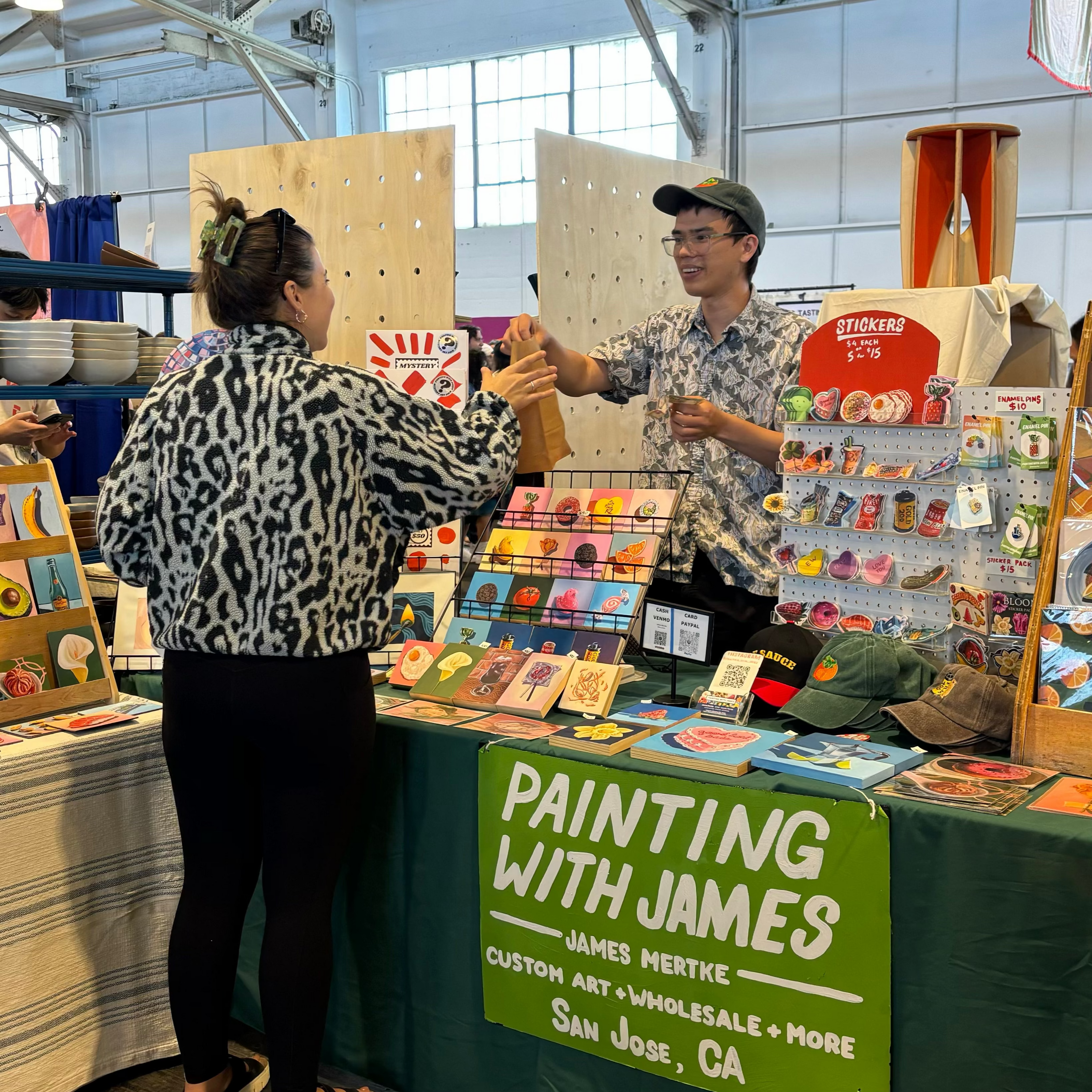 James Mertke stands behind his booth full of artwork and hands a customer wearing a black a white jacket a brown bag full of her purchases.