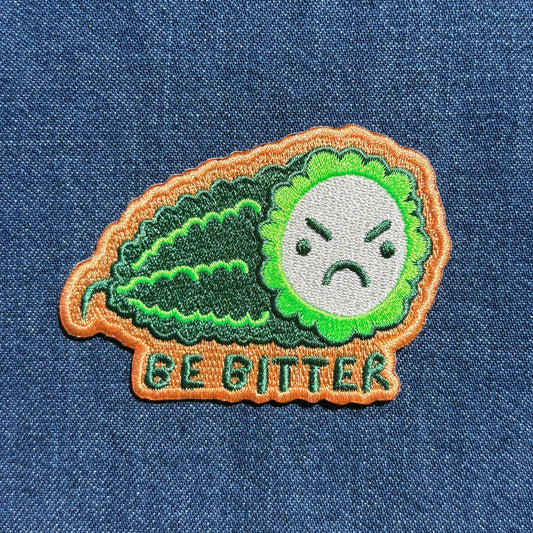 An embroidered patch shaped like a green bitter melon with an angry face and the text 'BE BITTER' below it, placed on a blue denim background.