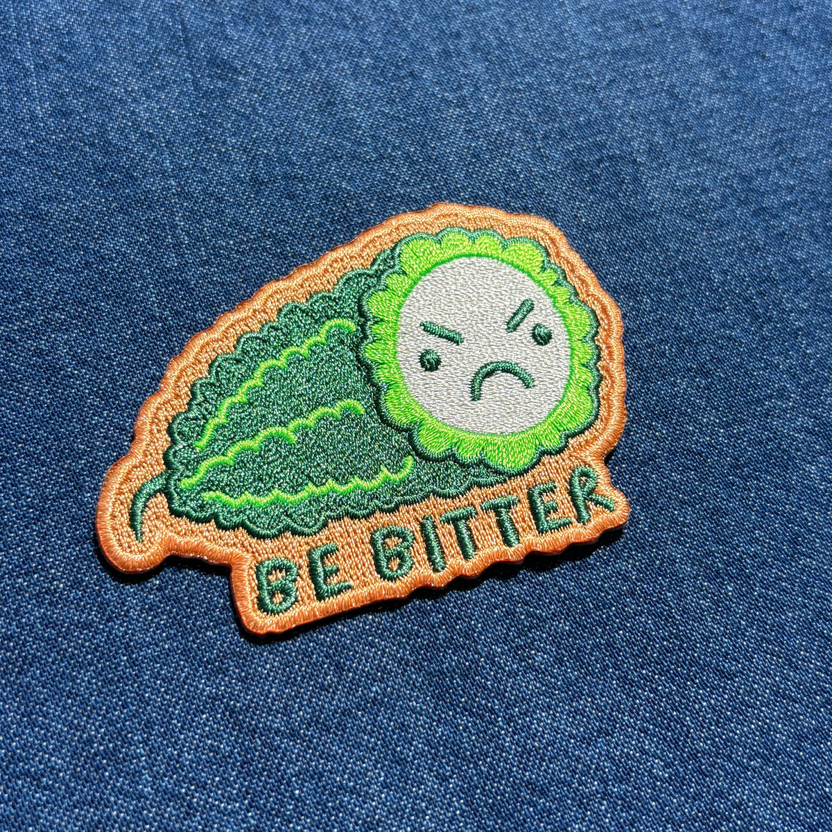 A close-up of an embroidered patch shaped like a green bitter melon with a sad face and the words 'BE BITTER' on a blue denim fabric background.