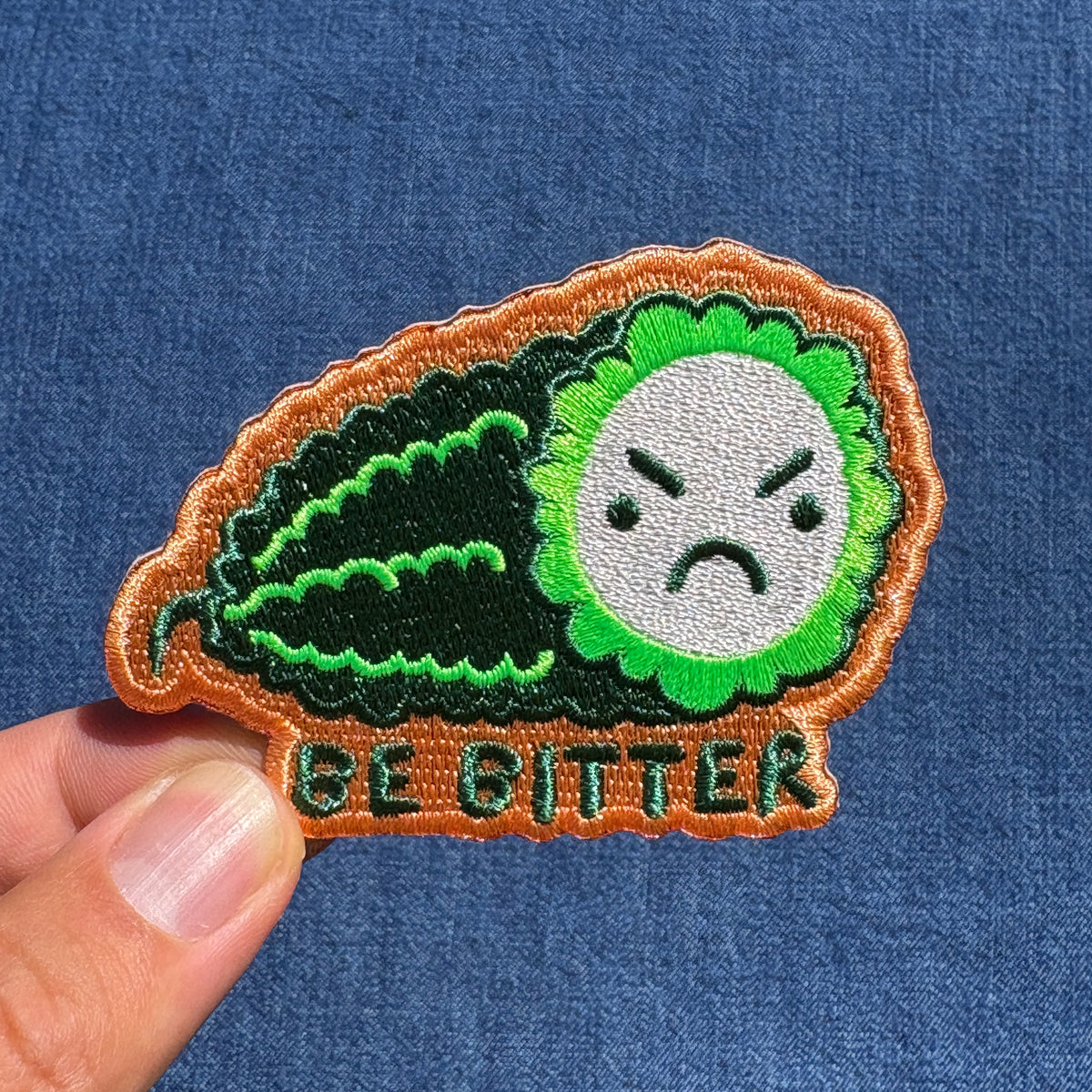 An embroidered patch shaped like a green bitter melon with an angry face and the text 'BE BITTER' at the bottom, held by a person's fingers against a blue denim background.