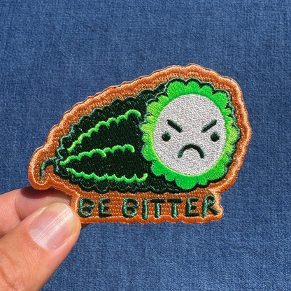 An embroidered patch shaped like a green bitter melon with an angry face and the text 'BE BITTER' at the bottom, held by a person's fingers against a blue denim background.