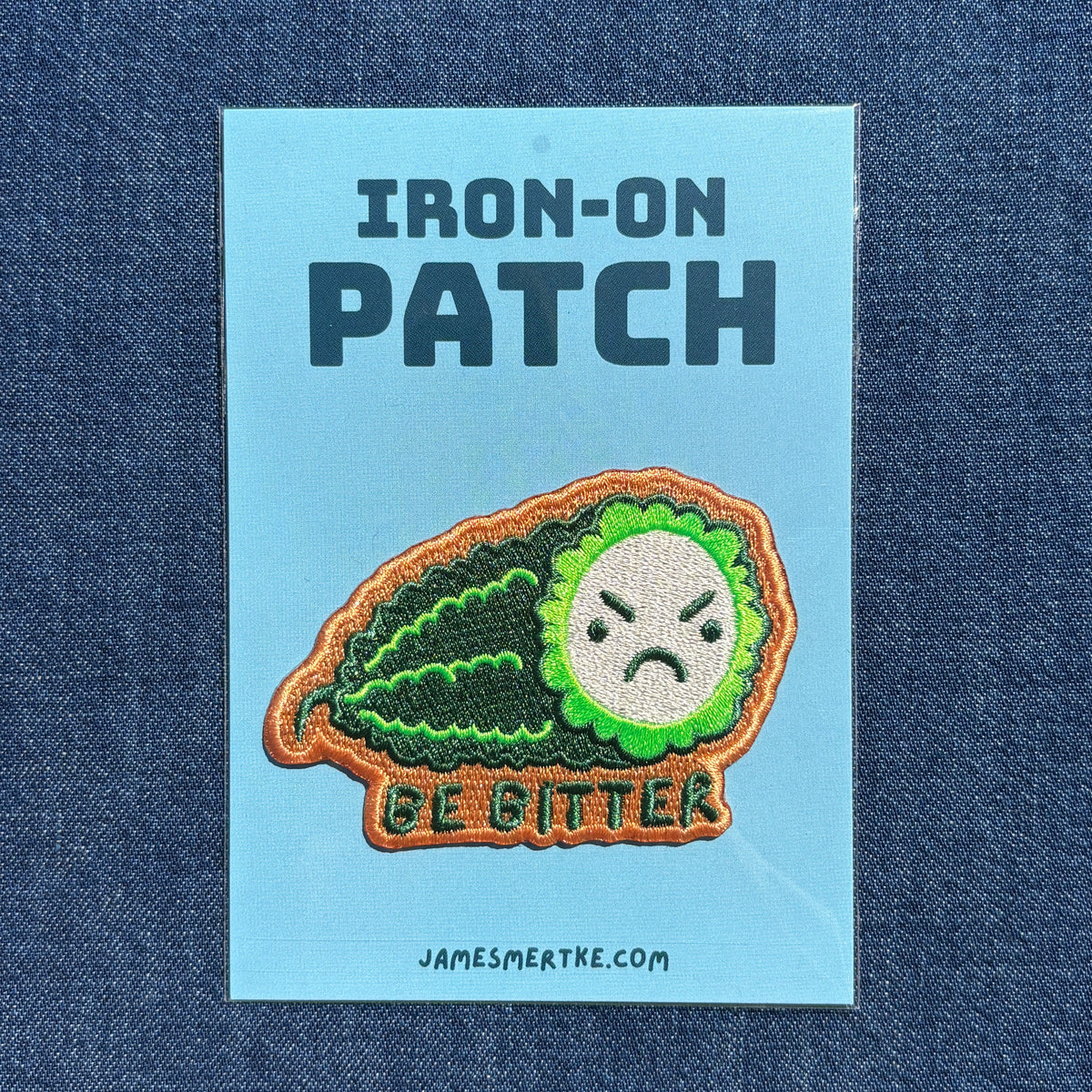 An iron-on patch featuring a green bitter melon with a frowning face and the words 'BE BITTER', displayed on a light blue backing card with the text 'IRON-ON PATCH' and 'JAMESMERTKE.COM', set against a dark blue denim background.