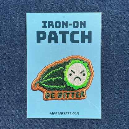 An iron-on patch featuring a green bitter melon with a frowning face and the words 'BE BITTER', displayed on a light blue backing card with the text 'IRON-ON PATCH' and 'JAMESMERTKE.COM', set against a dark blue denim background.