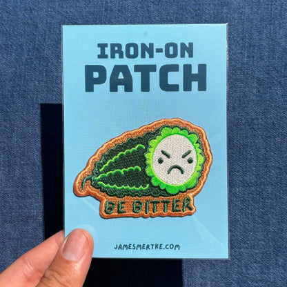 An embroidered iron-on patch featuring a green bitter melon with an angry face and the text 'BE BITTER' on a light blue backing card, held by a hand against a blue denim background.