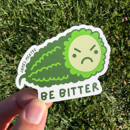 This sticker features an illustrated piece of  bitter melon with a grumpy face on its cross section. The phrase "BE BITTER" is written along the bottom. It is held in a hand.