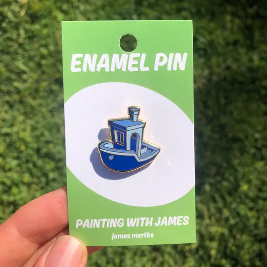 A blue boat enamel pin is attached to a green backing card. The boat is inspired by "Benchy", a common calibration print used to set up 3D printers.