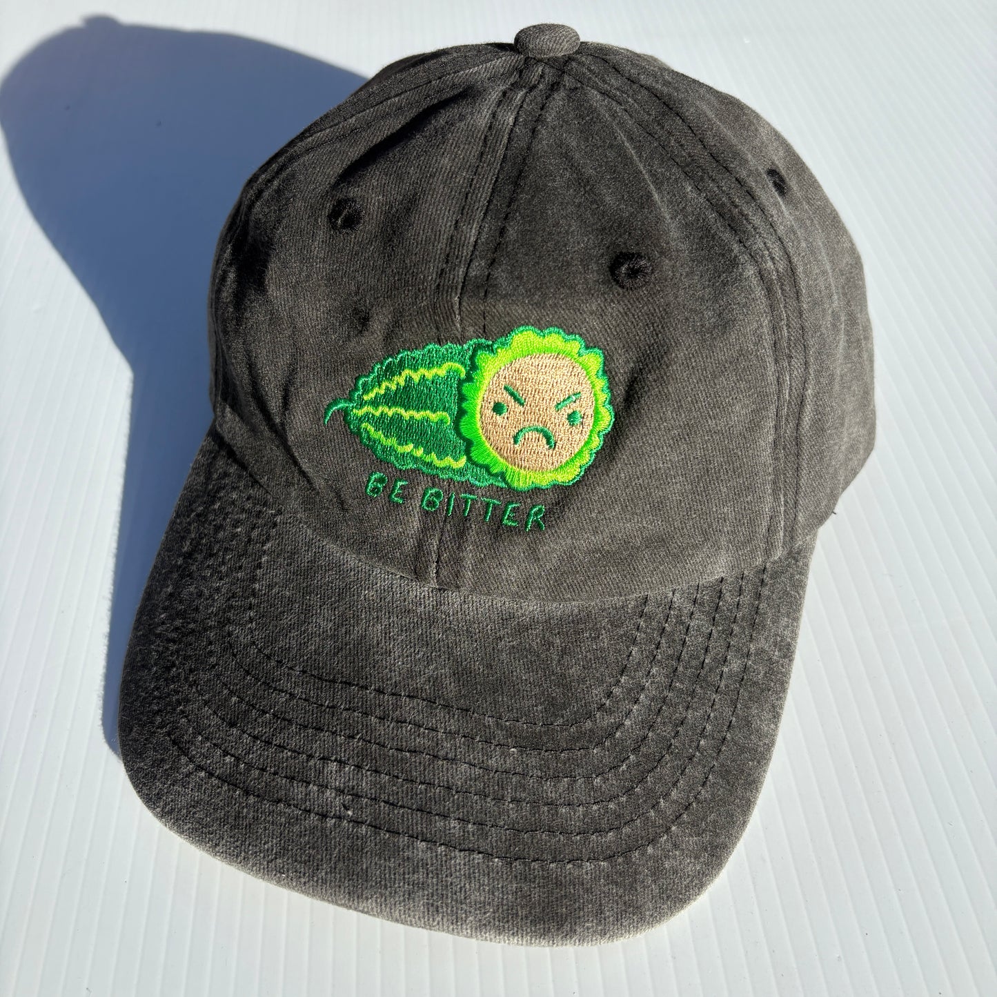 A black hat is embroidered with a green bitter melon design that has an angry face. The phrase "Be Bitter" is embroidered underneath it.