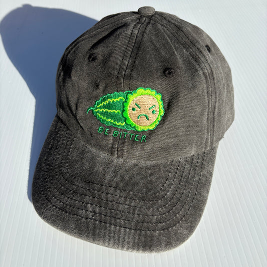 A black hat is embroidered with a green bitter melon design that has an angry face. The phrase "Be Bitter" is embroidered underneath it.