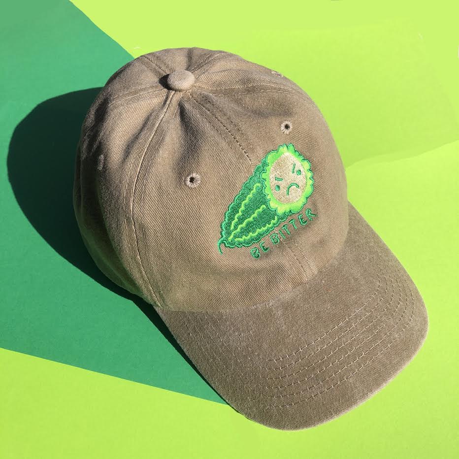 A beige hat is embroidered with a green bitter melon design that has an angry face. The phrase "Be Bitter" is embroidered underneath it.