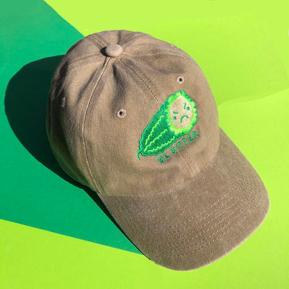 A beige hat is embroidered with a green bitter melon design that has an angry face. The phrase "Be Bitter" is embroidered underneath it.