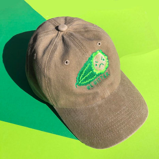 A beige hat is embroidered with a green bitter melon design that has an angry face. The phrase "Be Bitter" is embroidered underneath it.