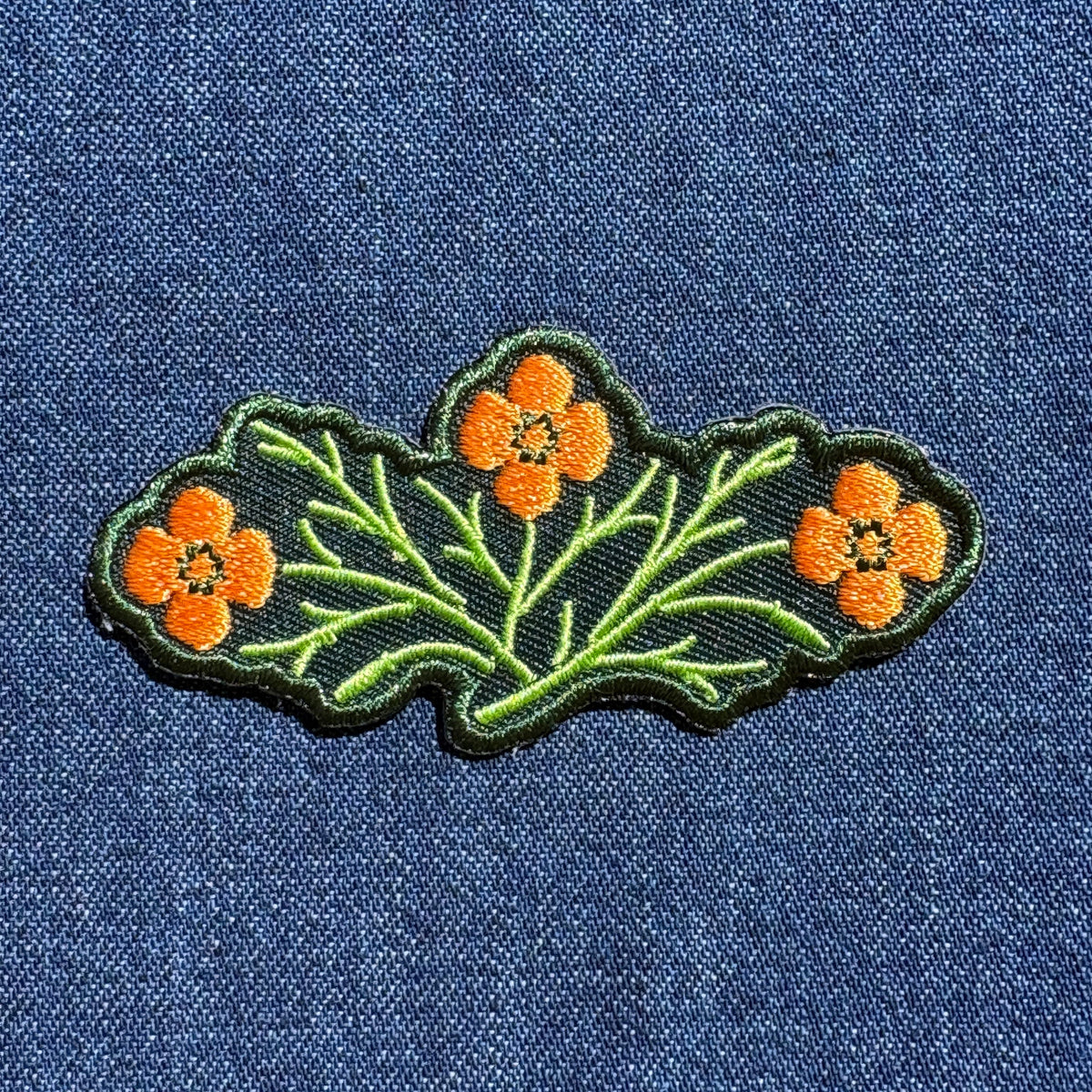 An embroidered patch featuring three orange flowers with green stems and leaves on a dark green background, shown on a blue denim fabric.
