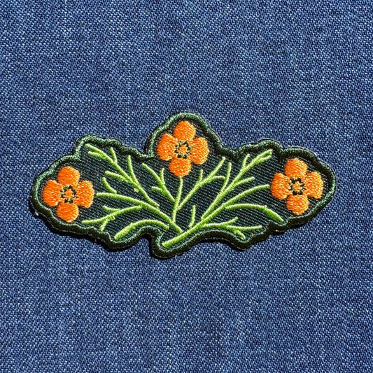 An embroidered patch featuring three orange flowers with green stems and leaves on a dark green background, shown on a blue denim fabric.