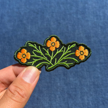 An embroidered patch featuring three orange flowers with green stems on a green background, held by a hand against a blue textured fabric backdrop.