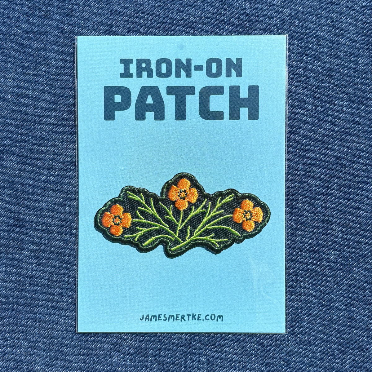 An iron-on patch featuring three orange flowers with green leaves on a green embroidered background, presented on a light blue backing card labeled 'IRON-ON PATCH' with the website JAMESMERTKE.COM, set against a blue denim fabric background.