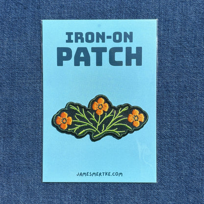 An iron-on patch featuring three orange flowers with green leaves on a green embroidered background, presented on a light blue backing card labeled 'IRON-ON PATCH' with the website JAMESMERTKE.COM, set against a blue denim fabric background.
