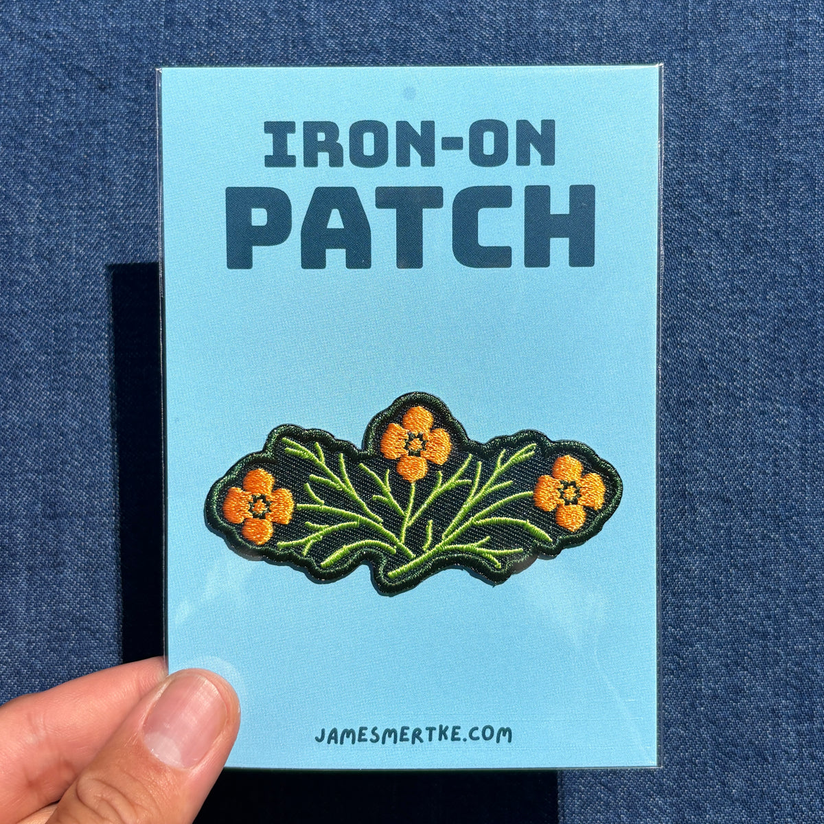 A hand holds a light blue backing card for an embroidered patch featuring California poppies. The card reads 'IRON-ON PATCH' at the top and 'JAMESMERTKE.COM.