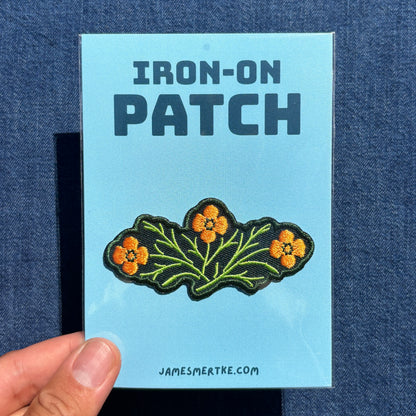 A hand holds a light blue backing card for an embroidered patch featuring California poppies. The card reads 'IRON-ON PATCH' at the top and 'JAMESMERTKE.COM.