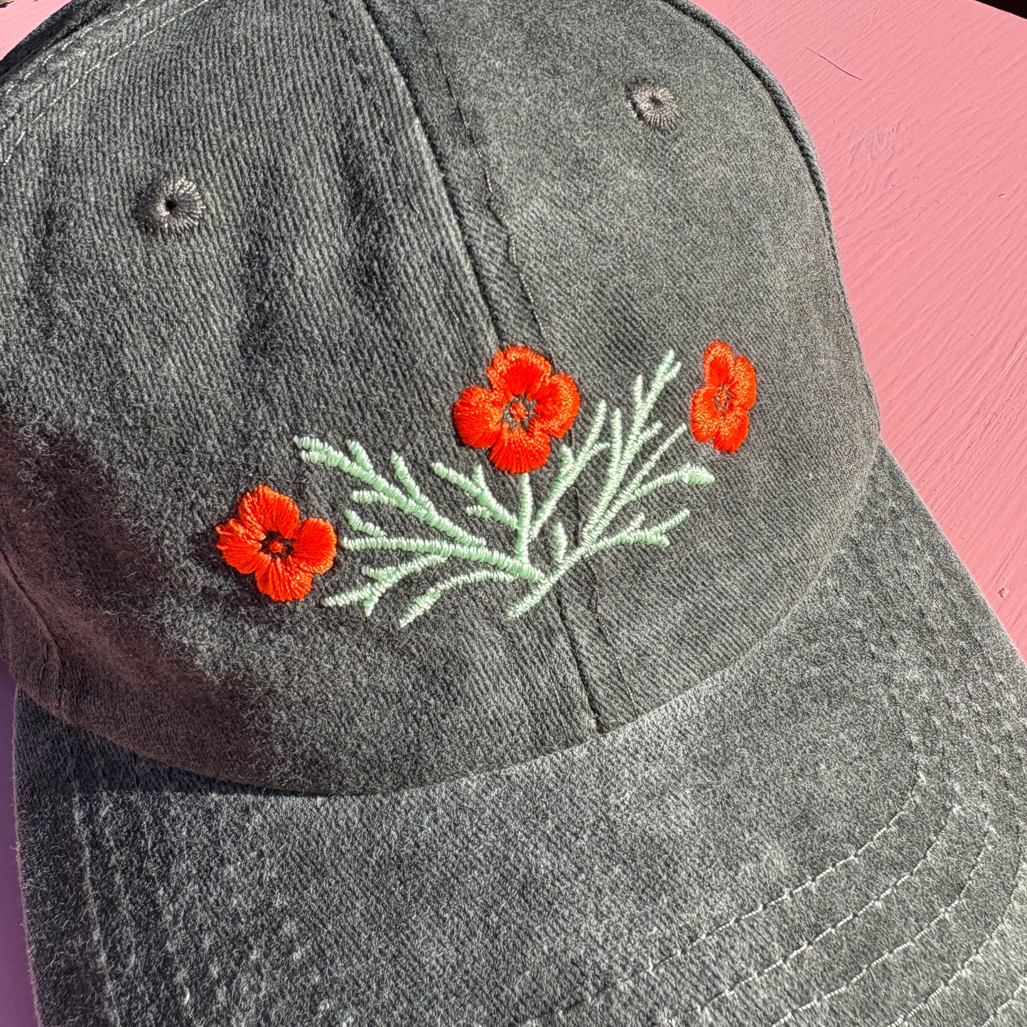 A close-up of a dark gray-green washed baseball cap featuring embroidered orange California poppy flowers and light green leaves on the front.