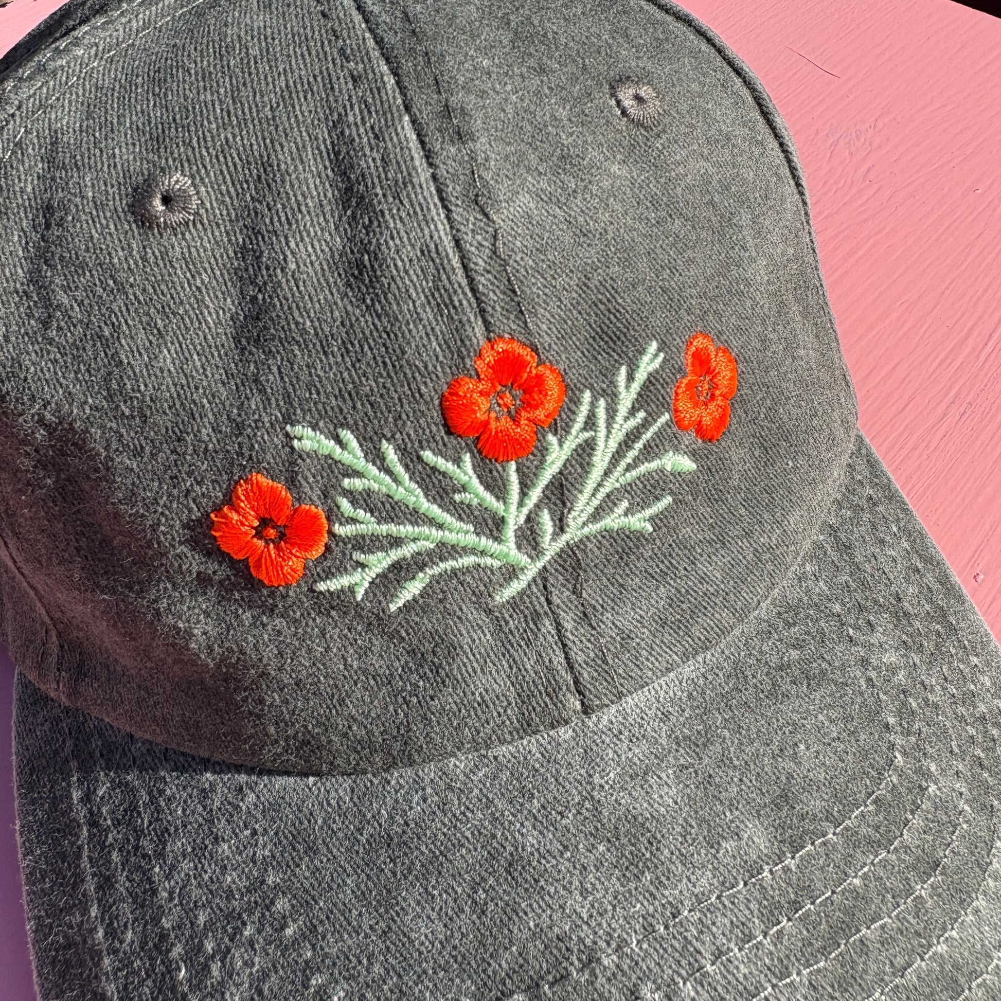 A close-up of a dark gray-green washed baseball cap featuring embroidered orange California poppy flowers and light green leaves on the front.