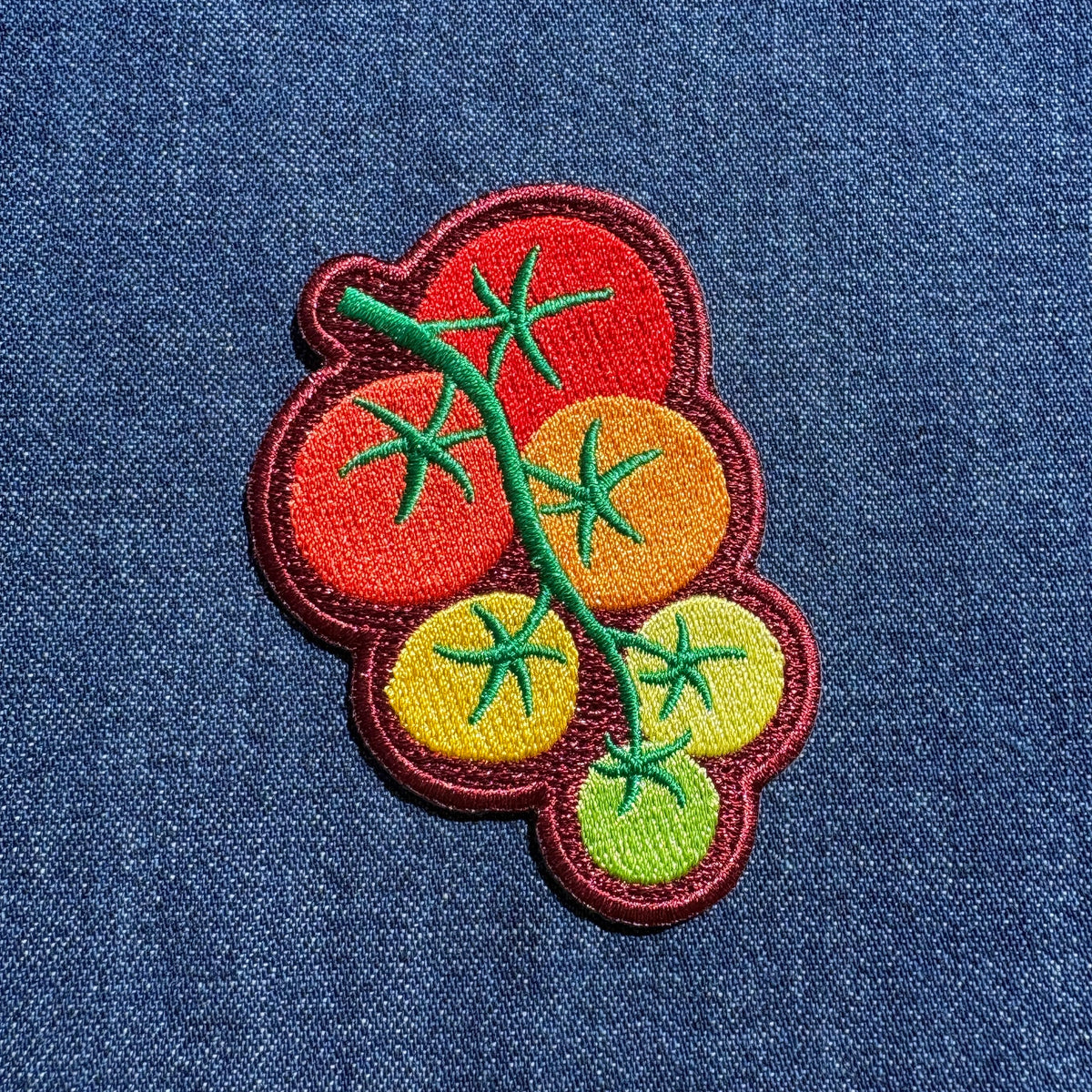 An embroidered patch of a tomato vine with red, orange, yellow, and green tomatoes on a blue denim fabric background.